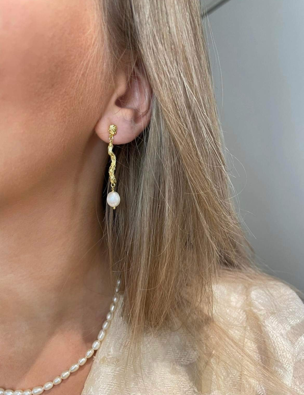Adalina guld øreringe hænger smukt i øret med sin ørestik funktion. Et organisk mellemled forbinder ørestikken og den ovale ferskvandsperle. En smuk værtindegave eller smykkegave.