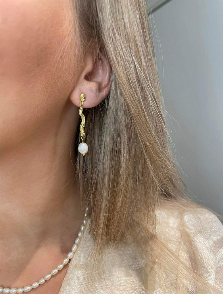 Adalina guld øreringe hænger smukt i øret med sin ørestik funktion. Et organisk mellemled forbinder ørestikken og den ovale ferskvandsperle. En smuk værtindegave eller smykkegave.